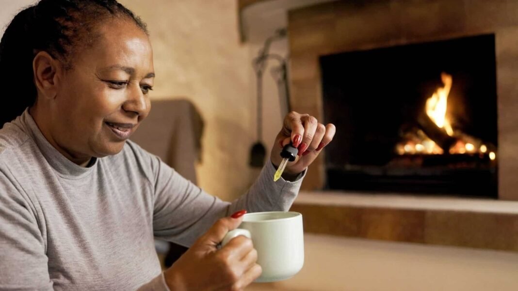 Cbd anxiety treatment – African senior woman taking cannabis oil in tea cup by cozy fireplace at home